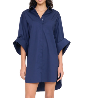 A woman wears the Staud Nico Dress, a loose-fitting navy cotton poplin mini shirtdress with adjustable rolled sleeves and a chest pocket, standing against a plain background.
