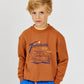 A young boy with blond hair wears the Arsene & Les Pipelettes Boys' Tropicana Sweatshirt, crafted from organic cotton and adorned with a car graphic and "Tropicana Surf Camp" print, paired with blue pants. He stands against a plain light background—an ideal option for comfortable casual wear.