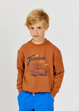 A young boy with blond hair wears the Arsene & Les Pipelettes Boys' Tropicana Sweatshirt, crafted from organic cotton and adorned with a car graphic and "Tropicana Surf Camp" print, paired with blue pants. He stands against a plain light background—an ideal option for comfortable casual wear.