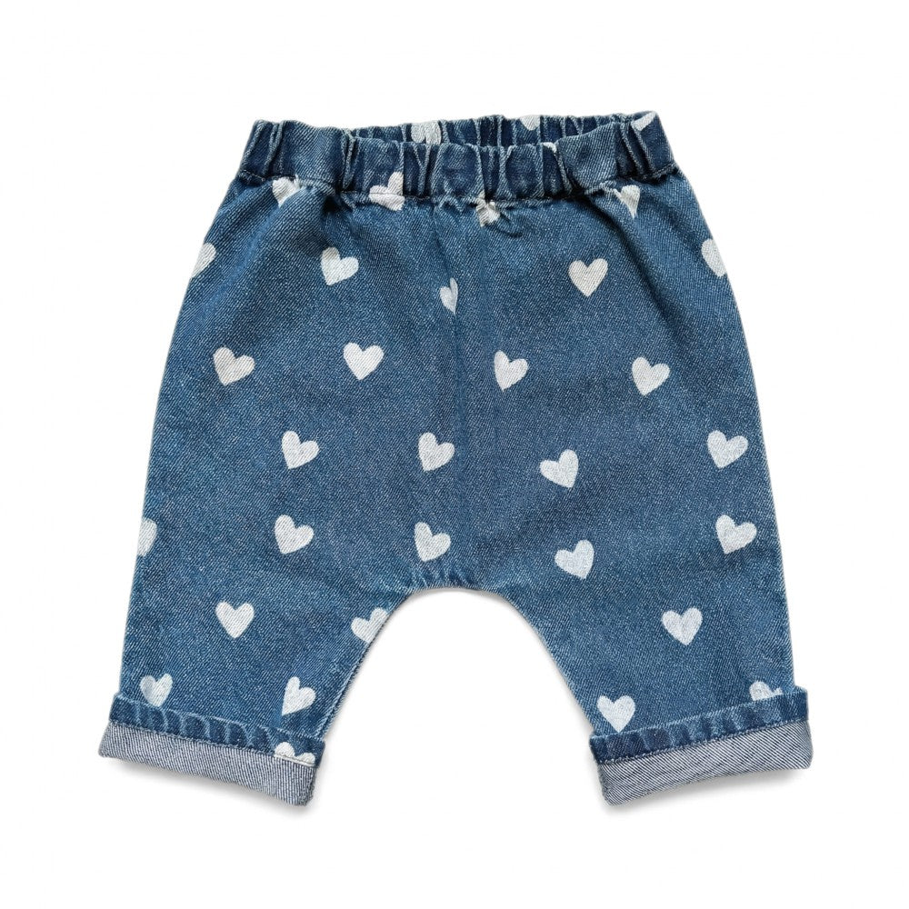 The Louis Louise Baby Girls’ Jungle Trouser features blue denim with an elastic waistband, a delightful white heart print, and rolled cuffs, displayed against a white background.