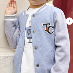 A young child wears the Tartine et Chocolat Baby Boys' and Boys' Easy Bomber Cardigan in light blue and gray with "T&C" patch lettering over a white graphic tee, standing indoors with one hand raised.