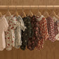A row of Rylee & Cru Baby Girls' Ada Rompers in floral and plaid designs hangs on wooden hangers against a wooden backdrop.