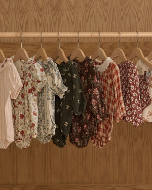 A row of Rylee & Cru Baby Girls' Ada Rompers in floral and plaid designs hangs on wooden hangers against a wooden backdrop.