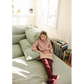 A young girl wearing the Girls’ Angele Shirt by Louis Louise and red tights sits on a light green couch near a window, with books stacked on the floor nearby.