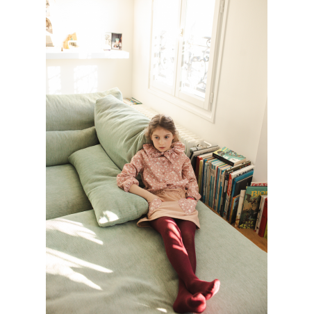 A young girl wearing the Girls’ Angele Shirt by Louis Louise and red tights sits on a light green couch near a window, with books stacked on the floor nearby.