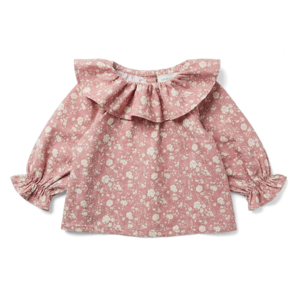 The Baby Girls’ Lilie Shirt by Louis Louise features a baby pink color with a white floral print, long sleeves, ruffled collar, and ruffled cuffs. The blouse is displayed laid flat on a white background.