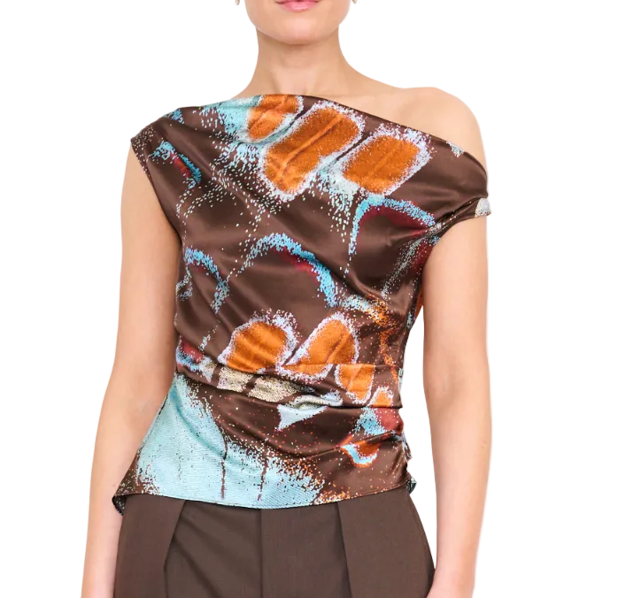 A woman wearing the Staud Phare Silk Top—a brown one-shoulder silk satin piece adorned with orange and blue abstract patterns and a drapey fitted silhouette—paired with brown pants.