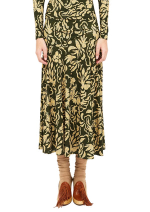 Wearing the Ulla Johnson Fiona Jersey Skirt—a long, dark green, high-waisted skirt with a gold floral print—paired with a matching long-sleeve top, beige socks, and brown fringe shoes; hands are relaxed at their sides.