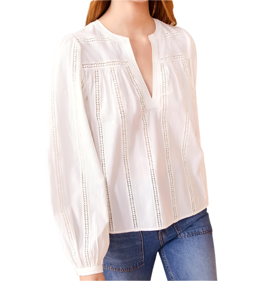 A person wears the Ulla Johnson Rowan Blouse in white, featuring intricate lace detailing and a V-neck, paired with blue jeans. The blouse is adorned with delicate geometric eyelets that add a touch of elegance.