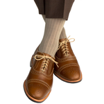 A person wearing brown dress shoes, brown pants, and Dapper Classics Taupe Solid Ribbed Mid Calf Socks with legs crossed against a white background.
