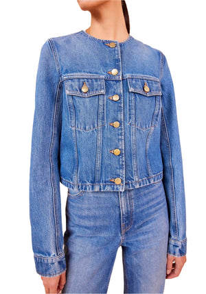 A person wearing the Ulla Johnson Sabine Jacket in a blue denim hue, accented with gold buttons and matching jeans, featuring a collarless neckline and vintage-inspired wash.