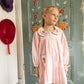 A young girl stands indoors near a decorated blue door, wearing the Louise Misha Baby Girls’ and Girls’ Ysee Dress in light pink cotton linen with delicate floral embroidery; two hats hanging on the wall beside her complement the hand-crocheted finishes on her charming attire.