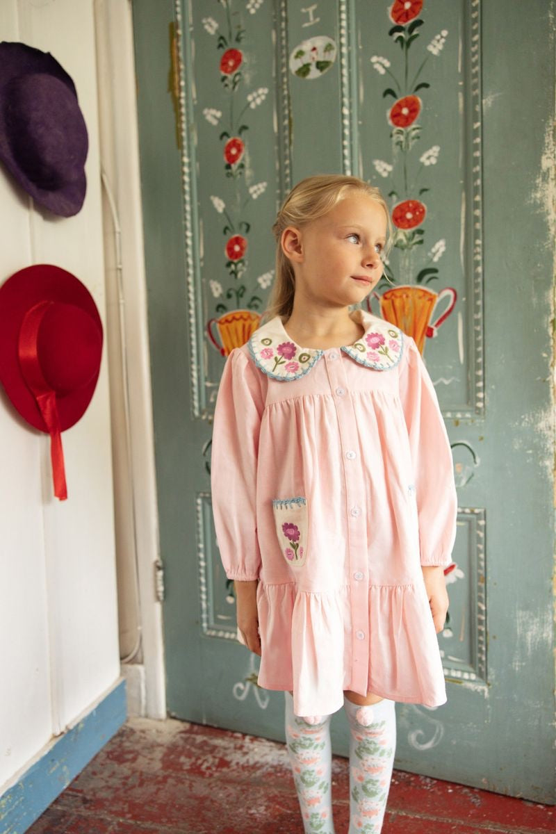 A young girl stands indoors near a decorated blue door, wearing the Louise Misha Baby Girls’ and Girls’ Ysee Dress in light pink cotton linen with delicate floral embroidery; two hats hanging on the wall beside her complement the hand-crocheted finishes on her charming attire.
