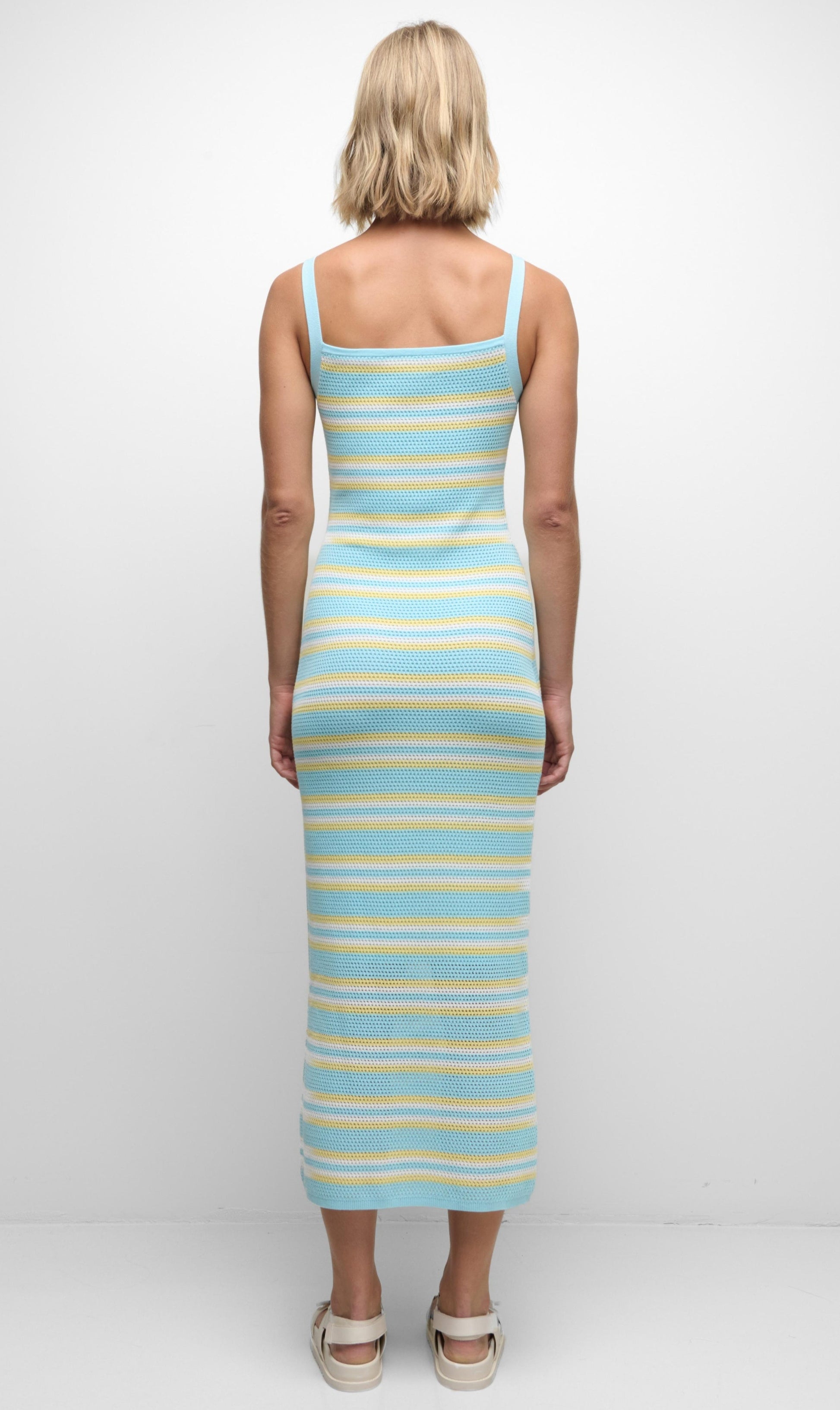 Person stands with back to camera, wearing the Simkhai Connelly Striped Midi Dress featuring blue, yellow, and white stripes, a ribbed square neckline, and slim fit, paired with white sandals against a plain white background.