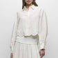 A woman wears the Simkhai Ames Linen Shirt—a long-sleeve, cream-colored piece with an embroidered floral hem—paired with a matching skirt; her face is partially out of frame.