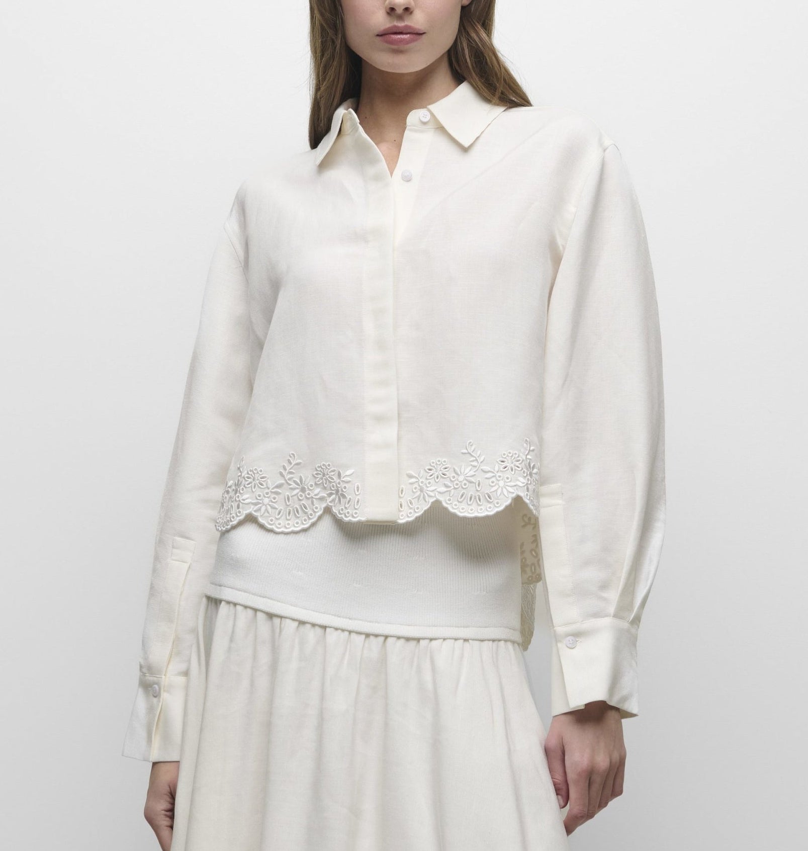 A woman wears the Simkhai Ames Linen Shirt—a long-sleeve, cream-colored piece with an embroidered floral hem—paired with a matching skirt; her face is partially out of frame.