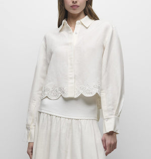A woman wears the Simkhai Ames Linen Shirt—a long-sleeve, cream-colored piece with an embroidered floral hem—paired with a matching skirt; her face is partially out of frame.