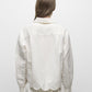 A person with long brown hair is shown from the back, wearing the Simkhai Ames Linen Shirt featuring a scalloped embroidered floral hem, paired with a matching white skirt.