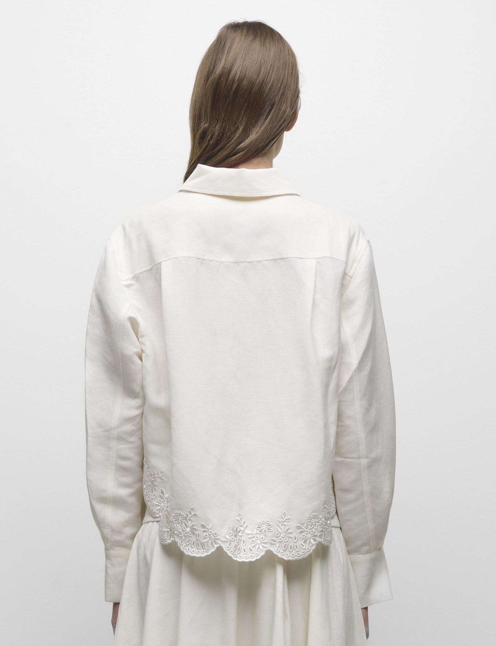A person with long brown hair is shown from the back, wearing the Simkhai Ames Linen Shirt featuring a scalloped embroidered floral hem, paired with a matching white skirt.