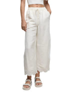 Model wears the Simkhai Ellison Linen Cropped Pant, featuring a scalloped hem, paired with a white crop top and sandals, standing against a white background.