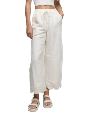 Model wears the Simkhai Ellison Linen Cropped Pant, featuring a scalloped hem, paired with a white crop top and sandals, standing against a white background.