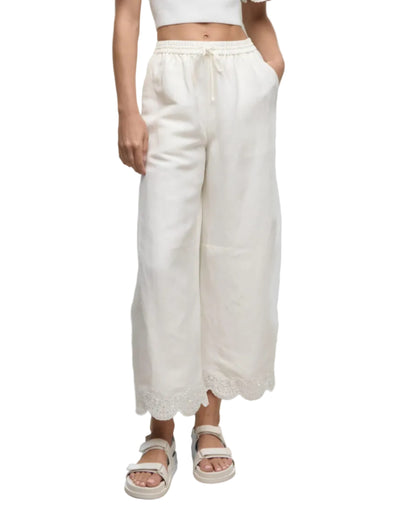 Model wears the Simkhai Ellison Linen Cropped Pant, featuring a scalloped hem, paired with a white crop top and sandals, standing against a white background.