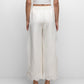 A person stands facing a white wall, wearing Simkhai's Ellison Linen Cropped Pant in cream with a scalloped hem, a matching short-sleeve top, and white sandals.