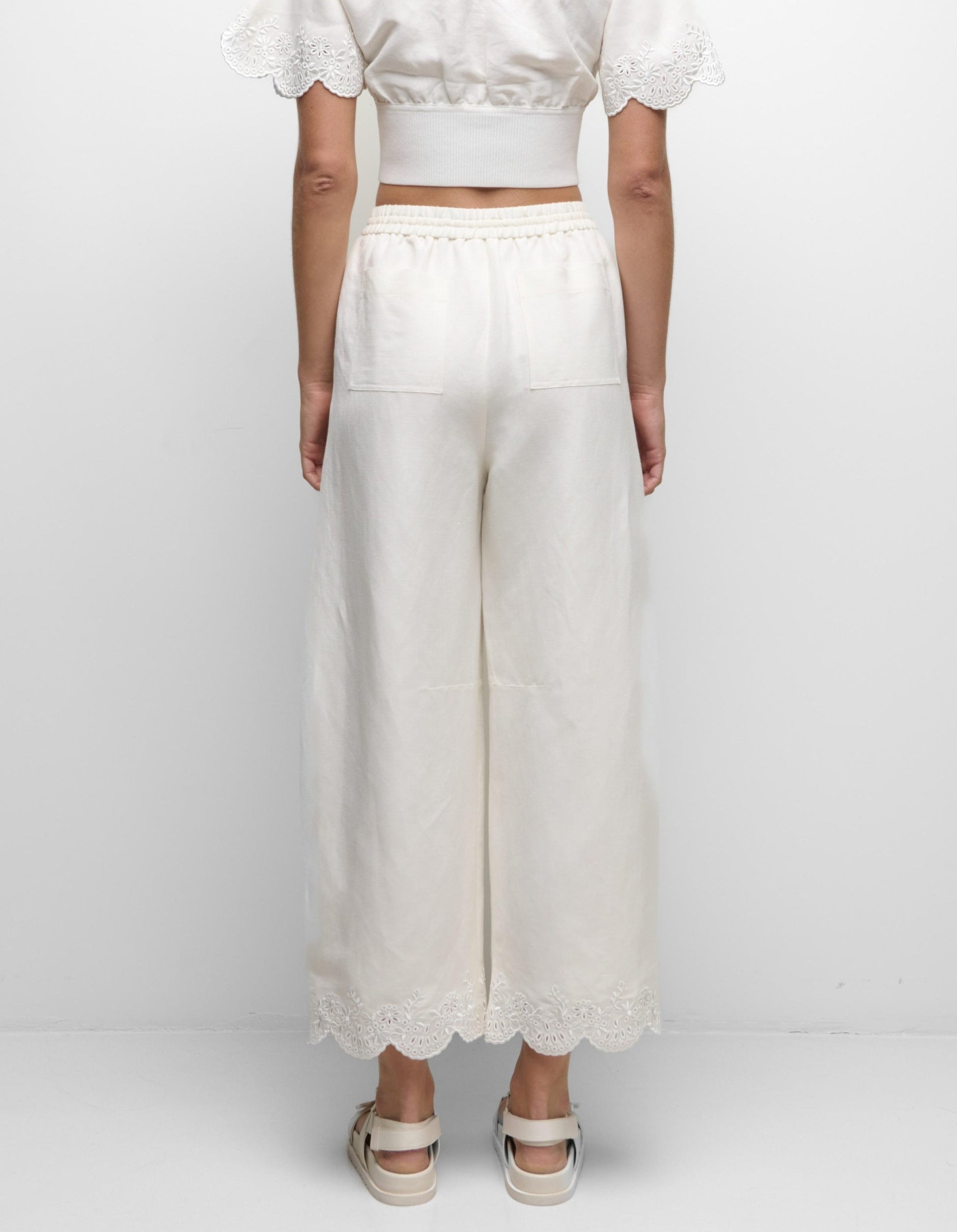 A person stands facing a white wall, wearing Simkhai's Ellison Linen Cropped Pant in cream with a scalloped hem, a matching short-sleeve top, and white sandals.