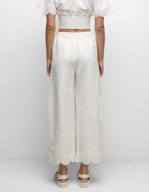 A person stands facing a white wall, wearing Simkhai's Ellison Linen Cropped Pant in cream with a scalloped hem, a matching short-sleeve top, and white sandals.