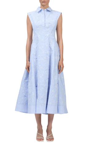 A woman in the Simkhai SK Neri Midi Dress—a sleeveless, light blue, collared dress with a fitted bodice and flared skirt—paired with open-toe heels.