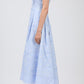 A person stands sideways wearing the Simkhai Neri Midi Dress—a light blue, sleeveless, collared dress with a subtle floral pattern—paired with white strappy heels.
