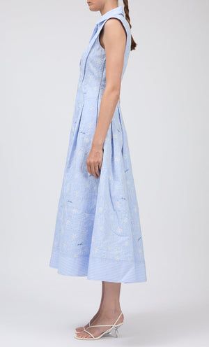 A person stands sideways wearing the Simkhai Neri Midi Dress—a light blue, sleeveless, collared dress with a subtle floral pattern—paired with white strappy heels.