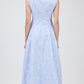 A woman faces away, wearing the Simkhai Neri Midi Dress in light blue with subtle floral pattern and a sleeveless, collared design, paired with white heeled sandals.