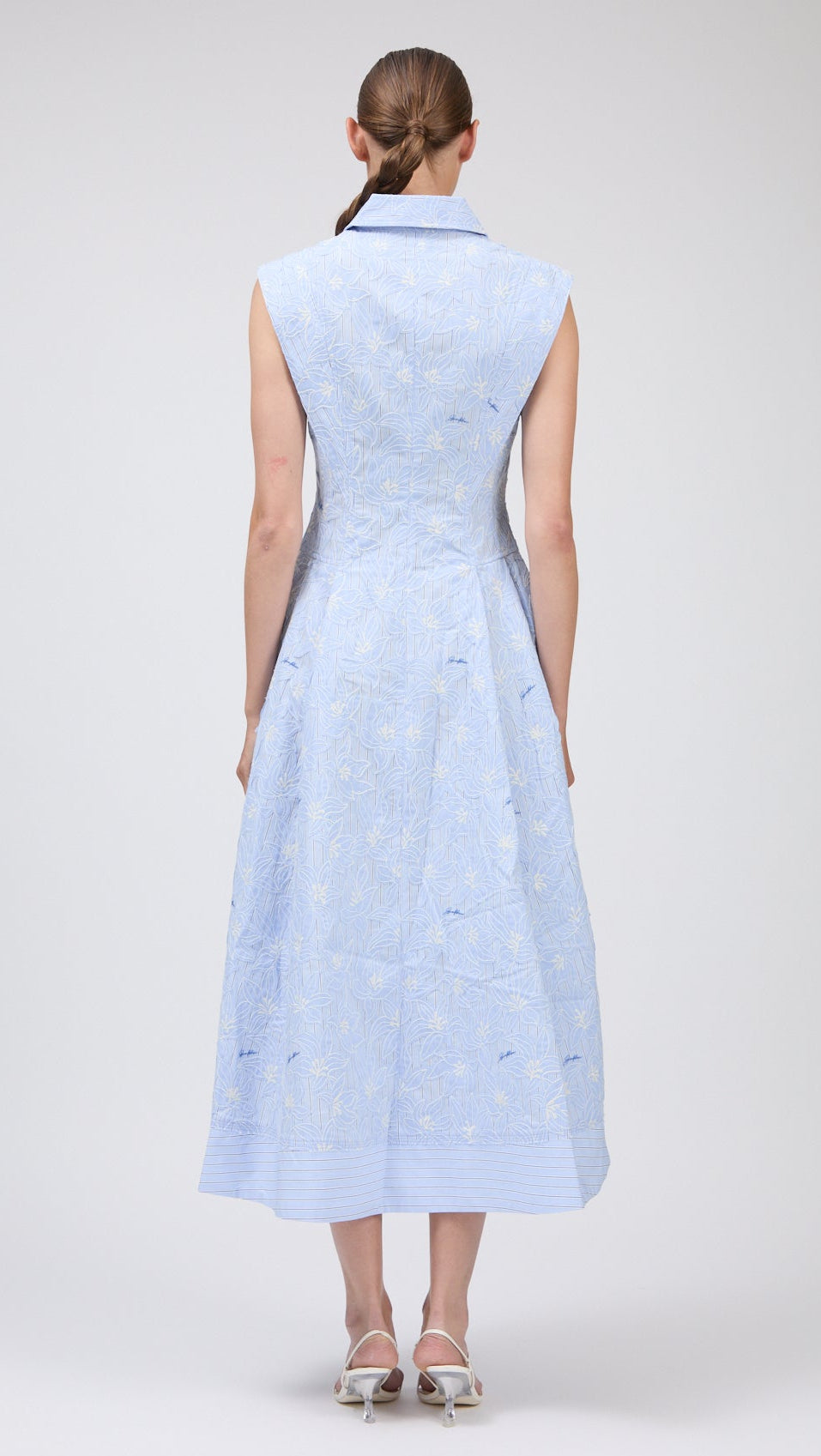 A woman faces away, wearing the Simkhai Neri Midi Dress in light blue with subtle floral pattern and a sleeveless, collared design, paired with white heeled sandals.