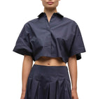 A person models the Simkhai Blair Shirt—navy blue, short-sleeve, cropped, button-up—paired with a matching high-waisted pleated skirt, shown from shoulders to mid-thigh against a white background.