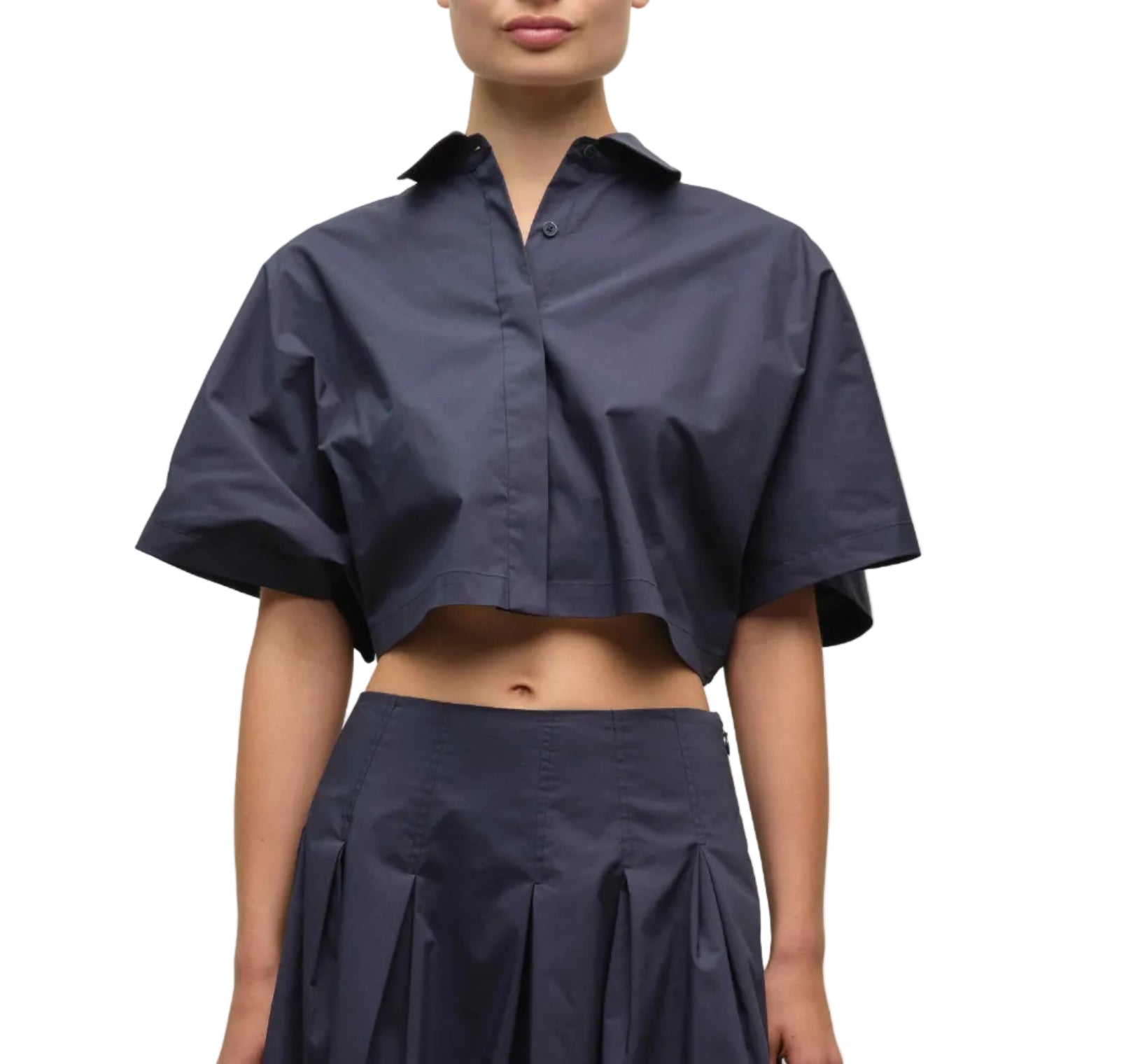 A person models the Simkhai Blair Shirt—navy blue, short-sleeve, cropped, button-up—paired with a matching high-waisted pleated skirt, shown from shoulders to mid-thigh against a white background.