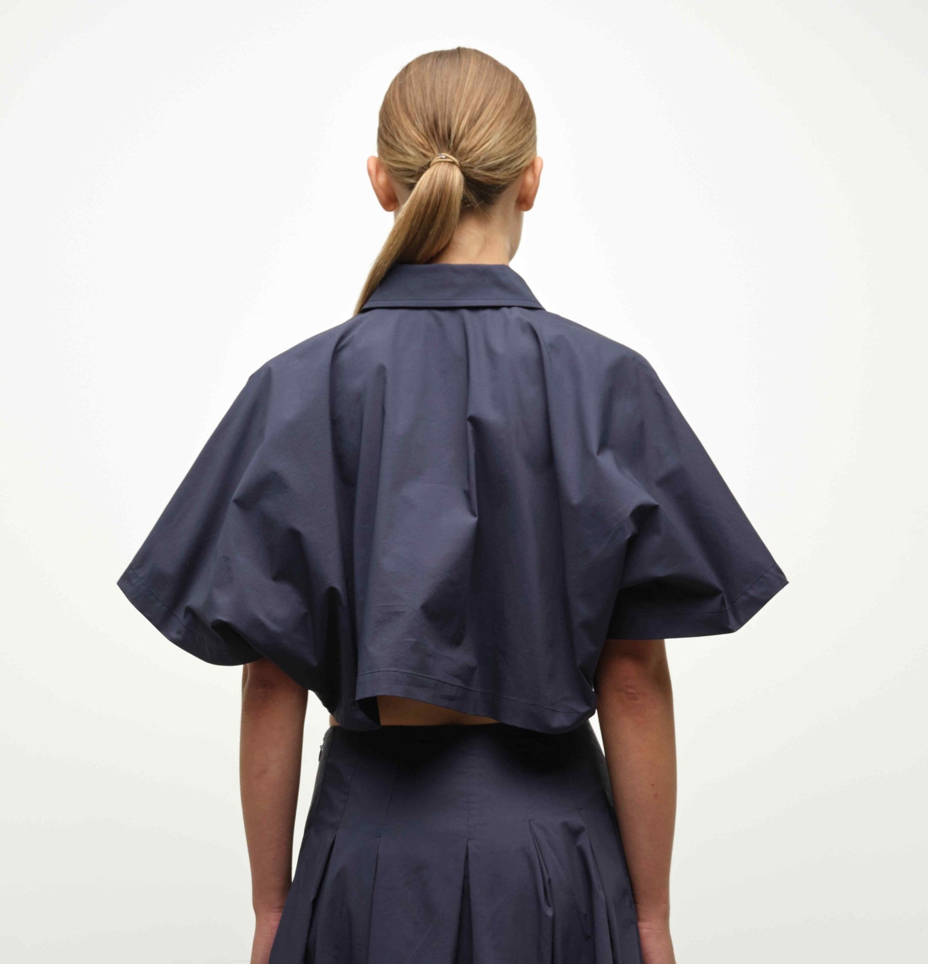 A person with straight, light brown hair in a low ponytail is seen from behind wearing the Simkhai Blair Shirt, a short-sleeved, dark navy cropped jacket, paired with a matching skirt against a plain background.