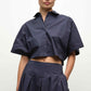 A person wears the dark blue, short-sleeved Blair Shirt by Simkhai, a cropped button-up, paired with matching pleated pants, standing against a plain background.