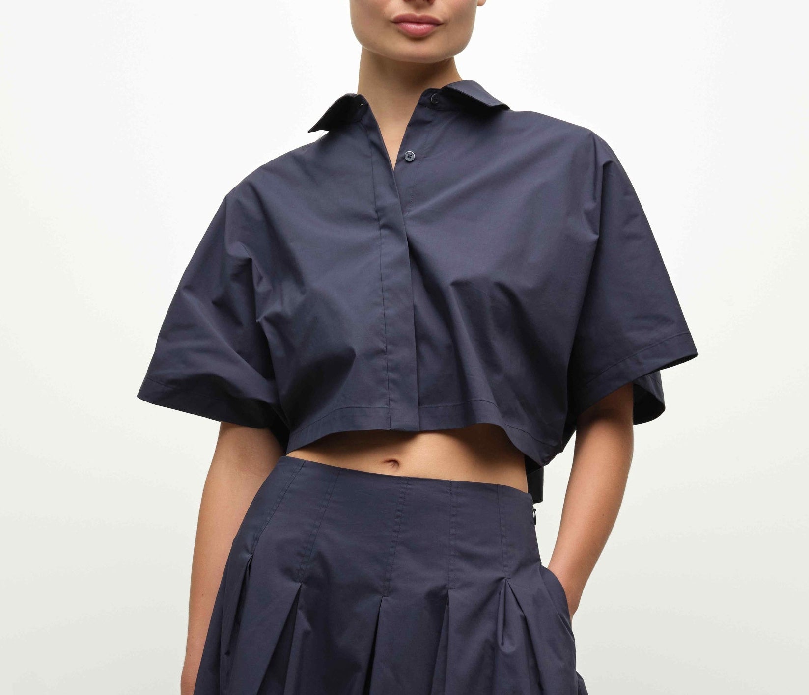 A person wears the dark blue, short-sleeved Blair Shirt by Simkhai, a cropped button-up, paired with matching pleated pants, standing against a plain background.