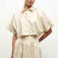 A person wearing the Simkhai Blair Shirt—a cream-colored, short-sleeve, cropped button-up—paired with a matching high-waisted skirt.