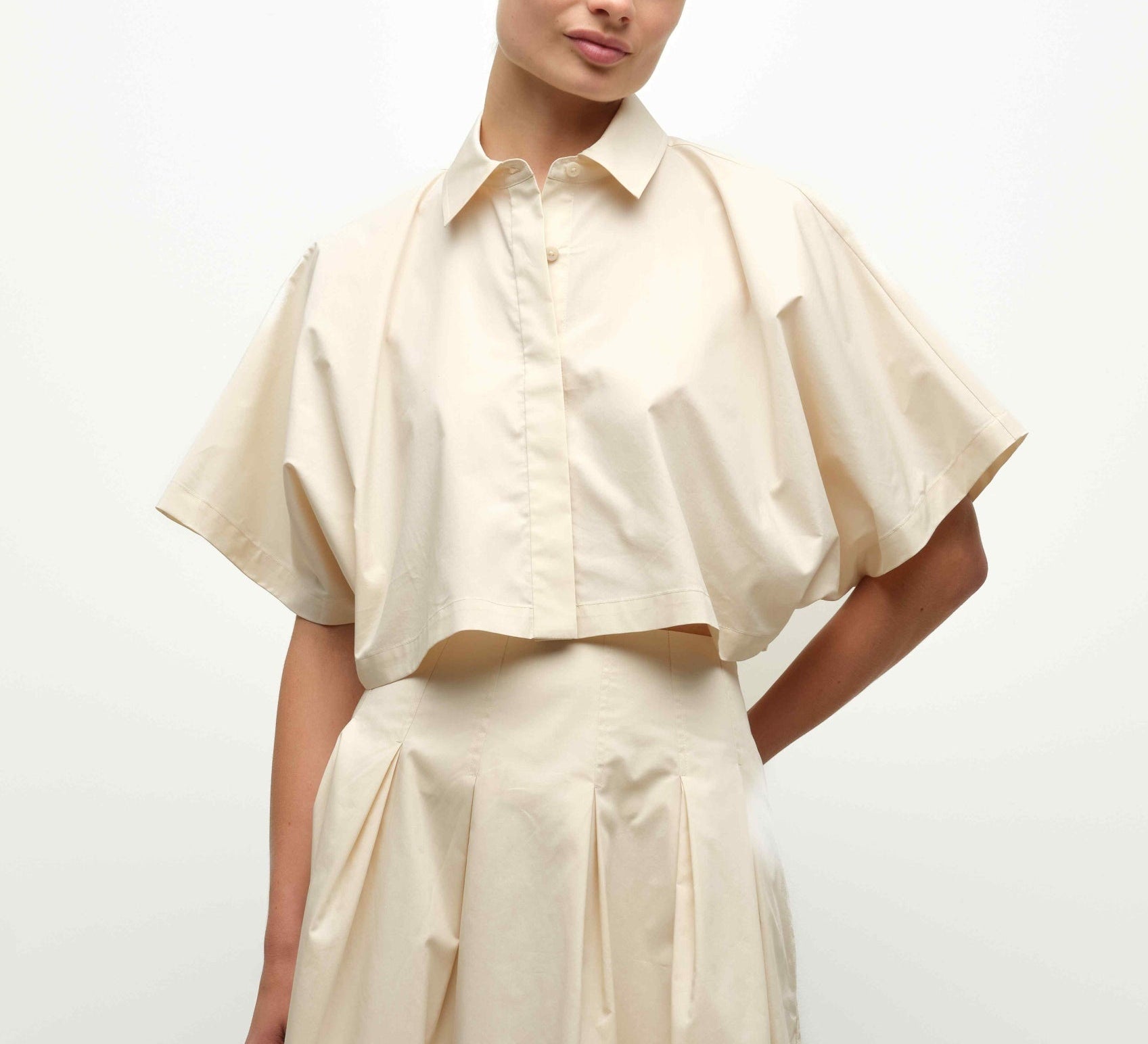 A person wearing the Simkhai Blair Shirt—a cream-colored, short-sleeve, cropped button-up—paired with a matching high-waisted skirt.
