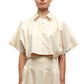 A woman wears the Simkhai Blair Shirt, a short-sleeve, cream-colored cropped button-up with a collar, paired with a high-waisted matching skirt.