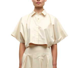 A woman wears the Simkhai Blair Shirt, a short-sleeve, cream-colored cropped button-up with a collar, paired with a high-waisted matching skirt.