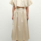 A person stands against a plain background wearing the Simkhai Elora Midi Skirt—a high-waisted, A-line, pleated beige skirt—paired with a light beige cropped button-up shirt and open-toe sandals.