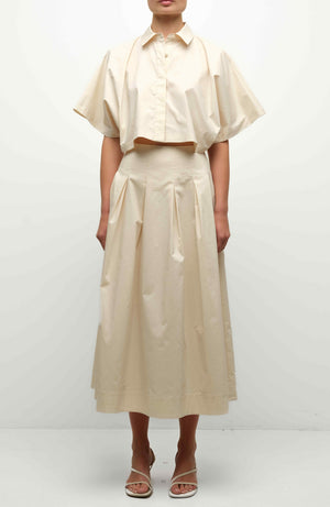 A person stands against a plain background wearing the Simkhai Elora Midi Skirt—a high-waisted, A-line, pleated beige skirt—paired with a light beige cropped button-up shirt and open-toe sandals.
