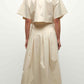 A woman stands facing away, wearing the Simkhai Elora Midi Skirt and a light beige short-sleeve cropped top with sandals. Her hair is tied back in a low ponytail.