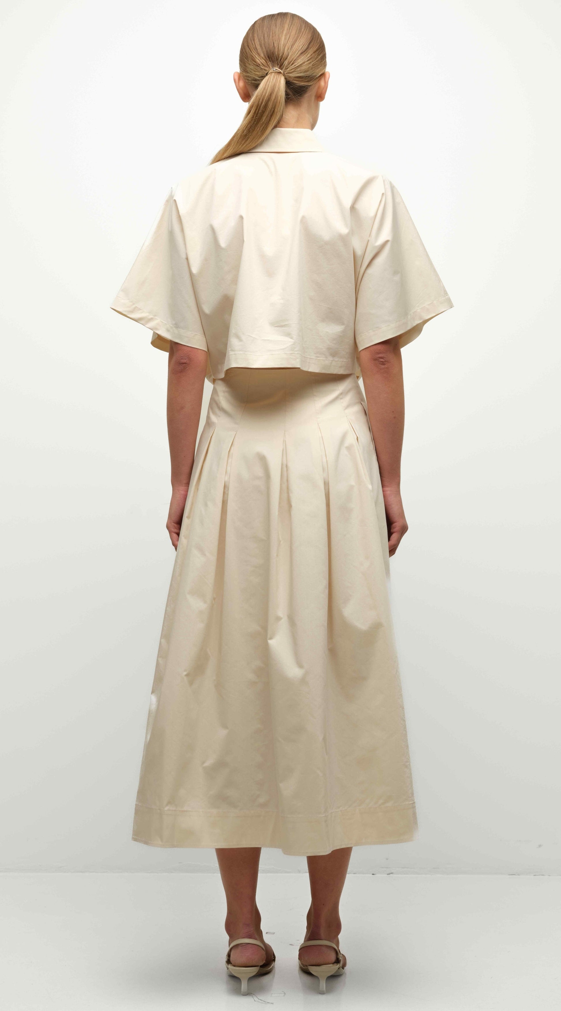 A woman stands facing away, wearing the Simkhai Elora Midi Skirt and a light beige short-sleeve cropped top with sandals. Her hair is tied back in a low ponytail.