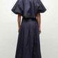 A woman faces away, wearing the Simkhai Elora Midi Skirt—a navy blue, pleated, midi-length skirt—with a matching short-sleeved cropped top and black heels.