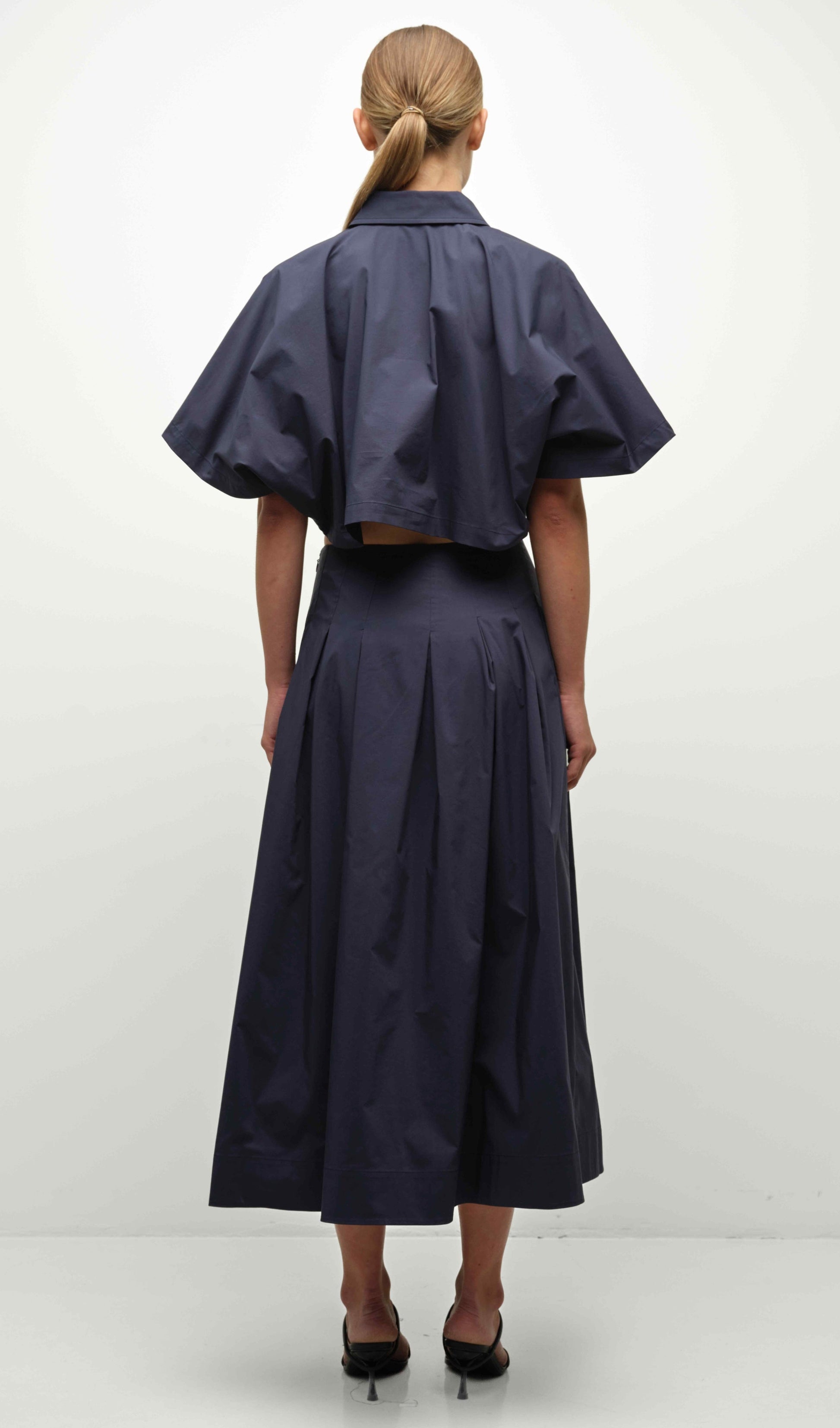 A woman faces away, wearing the Simkhai Elora Midi Skirt—a navy blue, pleated, midi-length skirt—with a matching short-sleeved cropped top and black heels.