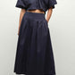 A person wears the Simkhai Elora Midi Skirt with a matching dark navy short-sleeve cropped button-up shirt and black sandals, standing against a plain background.